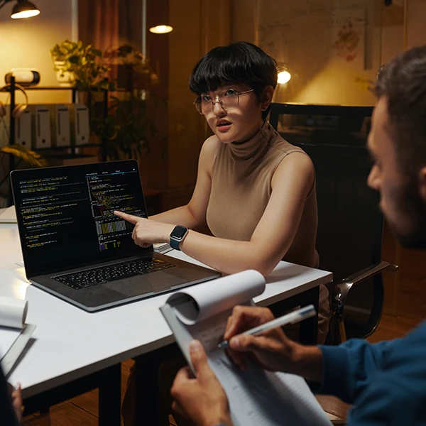 Mujer explicando código en la pantalla de una laptop a un colega durante una reunión de trabajo en oficina.