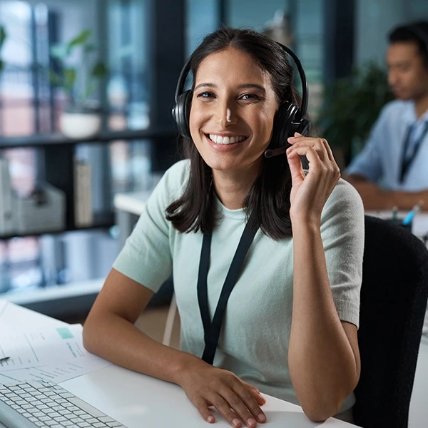 Mujer joven con auriculares y micrófono sonriendo mientras trabaja en atención al cliente en una oficina moderna.