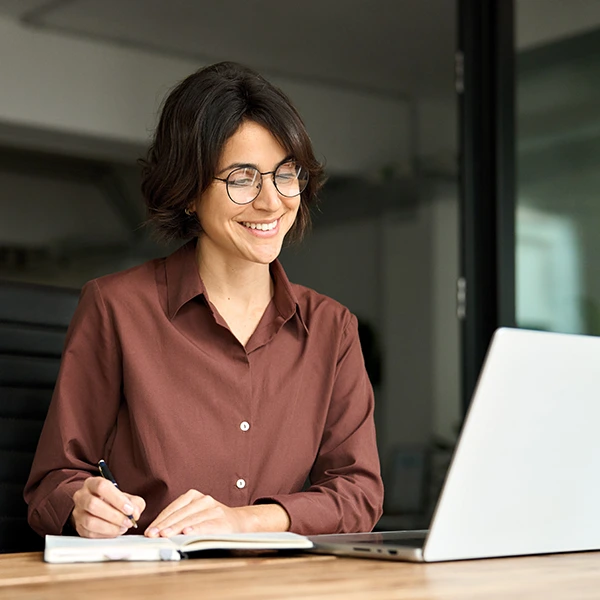 Mujer joven con gafas sonriendo mientras toma notas y trabaja en su laptop en una oficina.