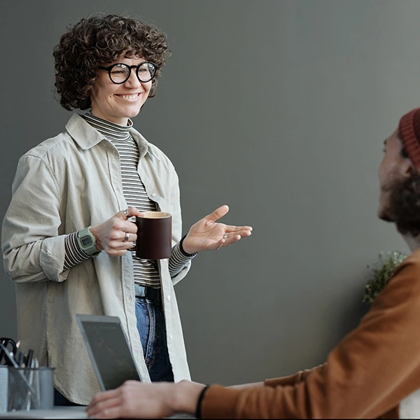 Mujer joven con gafas y cabello rizado sonriendo mientras sostiene una taza de café y conversa con un colega en una oficina.